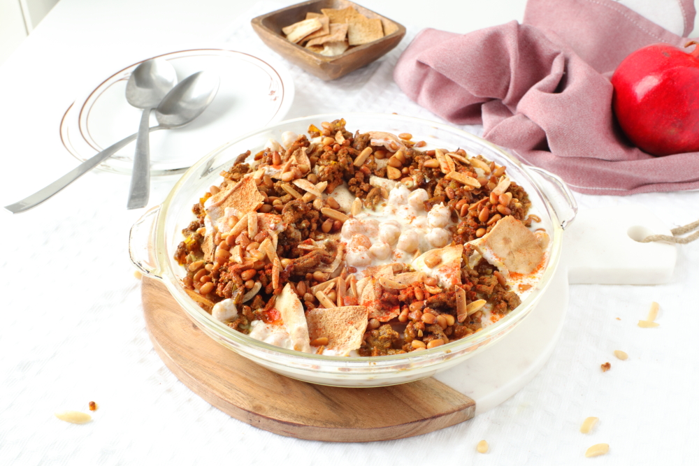 Fatteh meal dish on dinner table with cutlery, dishes, pita chips, and pomegranate